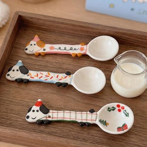 May include: Three ceramic spoons with dachshund dog designs. The spoons have white bowls and colourful dog figures. One dog has a pink and orange hat, another has a blue hat, and the third has a red hat. A small glass jug of milk is also present.