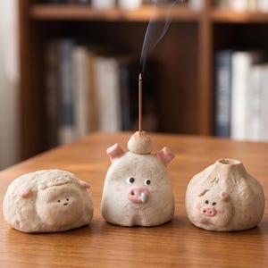 May include: Three small, textured, off-white ceramic figurines shaped like rounded animals. The center figure is a pig with pink ears and snout, holding a thin, lit incense stick. The left figure is a sheep, and the right figure is a piglet.