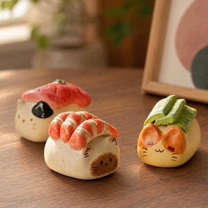 Może przedstawiać: Trzy małe ceramiczne figurki w kształcie kotów z dodatkami sushi. Jeden kot ma kawałek czerwonej ryby, drugi pomarańczowe krewetki, a trzeci zielone plasterki awokado. Każdy kot ma namalowane cechy twarzy, w tym oczy i wąsy.