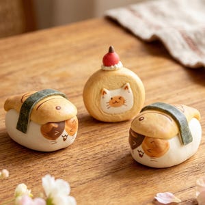 Op de afbeelding: Drie kleine decoratieve beeldjes in de vorm van etenswaren met kattengezichten. Twee beeldjes lijken op sushirolletjes met beige en witte lichamen en donkergroene zeewierstrips. Eén rond beeldje heeft een kattengezicht en een kleine rode bes.