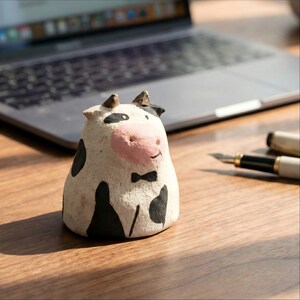 Pode incluir: Uma estatueta de vaca artesanal com base branca, manchas pretas e nariz rosa. A vaca tem pequenos chifres e uma gravata borboleta preta. A estatueta está sobre uma superfície de madeira, com um laptop e uma caneta no fundo.