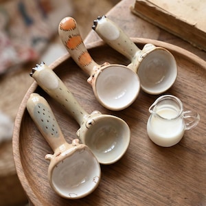 May include: Four ceramic spoons with animal designs, including bear and giraffe motifs, are displayed on a wooden tray. The spoons have beige handles and white bowls. A small glass creamer with milk is also present.