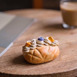 May include: A close-up shot of a pastry on a wooden surface. The pastry is oval-shaped with a golden-brown crust and a decorative pattern. It is topped with swirls of light-colored cream and garnished with a blue and yellow piece. A glass of coffee is in the background.