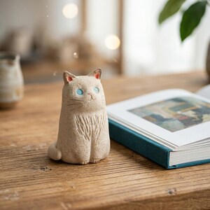 May include: A beige ceramic cat figurine with blue eyes and a carved chest detail. The cat has small, pointed ears with red accents. It sits upright on a wooden surface, near an open book with a teal cover.