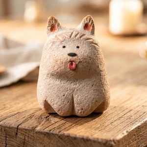 Op de afbeelding: Een beige keramieken hondenbeeldje met een getextureerd oppervlak. De hond heeft een zwarte neus, rode oren en een roze tong. Het beeldje staat op een houten tafel.