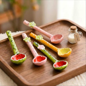 Pode incluir: Quatro colheres de cerâmica coloridas com tigelas com tema de frutas, incluindo desenhos de melancia, morango e limão. As colheres estão em uma bandeja de madeira, com um pequeno pote de creme. As colheres têm detalhes florais.