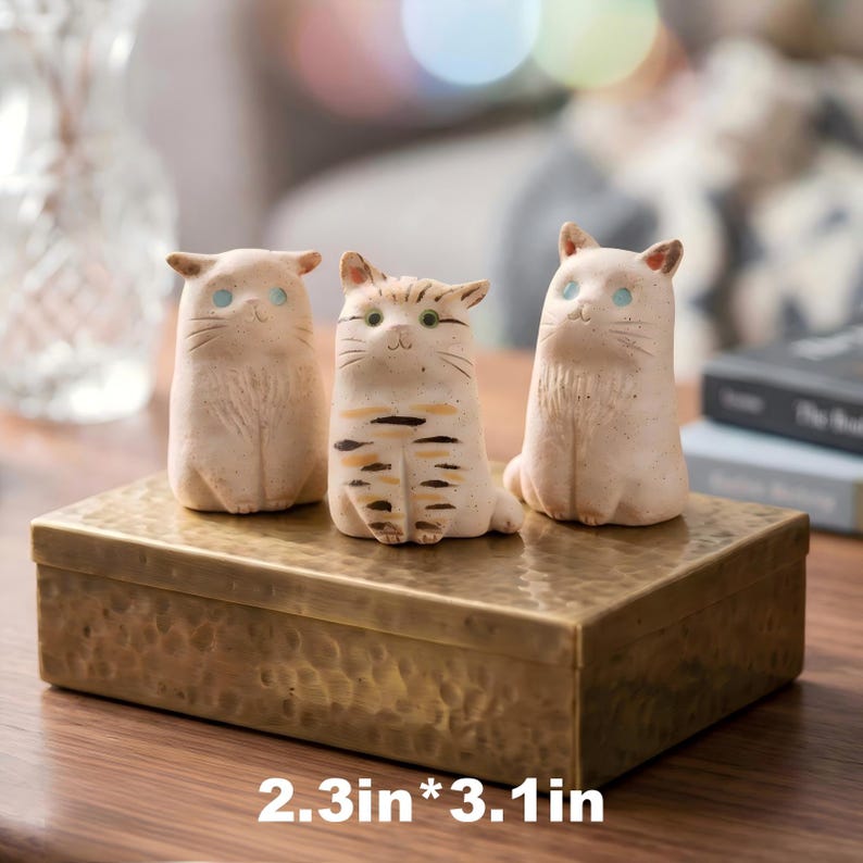 May include: Three ceramic cat figurines, each with unique markings and expressions, sit atop a textured gold-colored box. The cats are approximately 2.3 inches by 3.1 inches. The figurines have blue or green eyes and are in a neutral color palette.