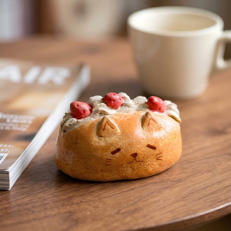 May include: A ceramic cat-shaped dessert with a strawberry topping. The orange-brown pastry has a cream filling and three red strawberry accents. The cat's face is detailed with small eyes, a nose, and whiskers. A white mug and magazine are in the background.