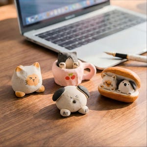 Könnte beinhalten: Eine Sammlung handgefertigter Keramik-Tierfiguren, darunter eine Katze, ein Hund und eine Katze in einer rosa Tasse mit Kirschdetails. Die Figuren befinden sich auf einer Holzoberfläche in der Nähe eines Laptops und eines Stifts.
