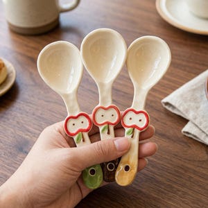 Może przedstawiać: Trzy ceramiczne łyżki z uchwytami w kształcie jabłek. Łyżki mają kremowe miseczki i są trzymane w dłoni. Wzory jabłek mają kolory czerwony, zielony i brązowy. Łyżki są prawdopodobnie przeznaczone do serwowania lub jedzenia.