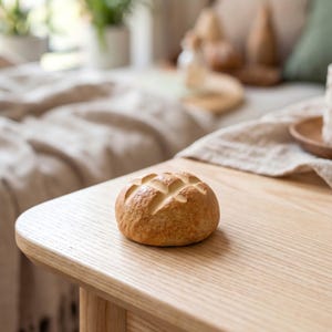 May include: A round, golden-brown bread roll with a cross-hatched pattern on top sits on a light-colored wooden table. The bread is the focal point, with a blurred background suggesting a domestic setting.