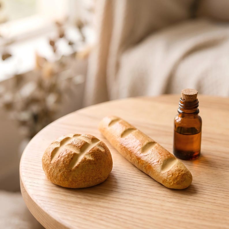 May include: A still-life image featuring a round bread roll and a baguette, both with a golden-brown crust, alongside a small amber glass bottle with a cork stopper. The items are arranged on a light-colored wooden table.