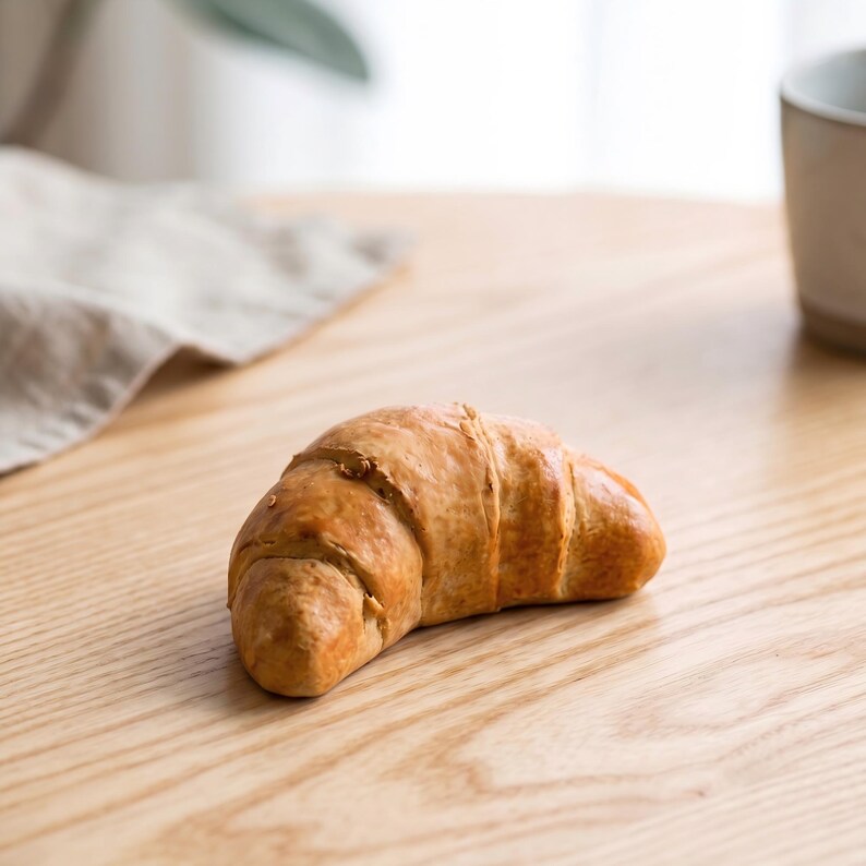 May include: A golden-brown croissant rests on a light-colored wooden surface. The pastry has a flaky texture and is partially in focus. A neutral-colored cloth and a ceramic mug are in the background, suggesting a breakfast or brunch setting.
