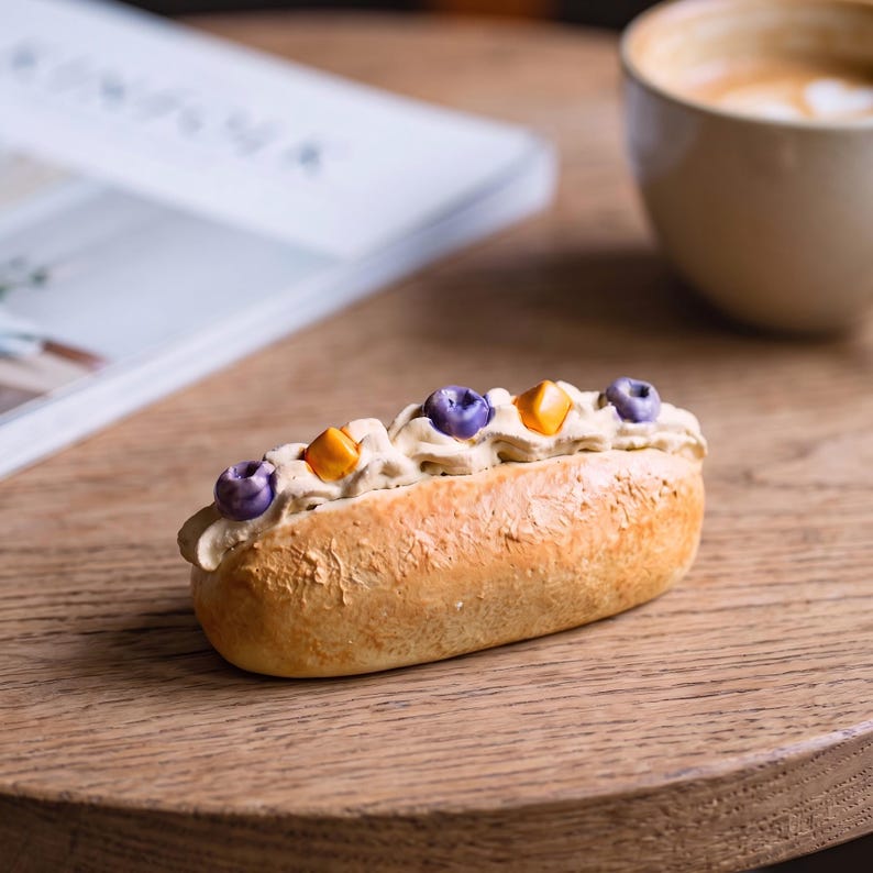 May include: A miniature eclair with a light brown exterior, topped with cream and decorated with purple and orange candies. The dessert sits on a wooden surface, with a coffee cup and a magazine in the background.