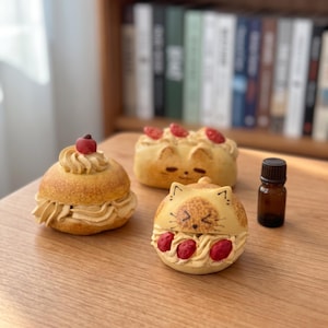 Könnte beinhalten: Drei dekorative Objekte in Gebäckform, darunter ein rundes, mit Creme gefülltes Brötchen mit einer roten Beere, ein rechteckiges Brötchen mit Katzengesicht und roten Beeren sowie ein rundes Brötchen mit Katzengesicht und roten Beeren neben einer braunen Glasflasche.