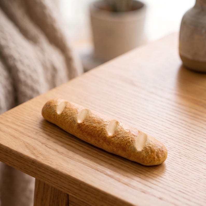 May include: A miniature baguette with a golden-brown crust and three diagonal cuts, resting on a light-colored wooden table. The bread's texture appears slightly rough, and the color gradient suggests baking. The scene is lit with natural light.