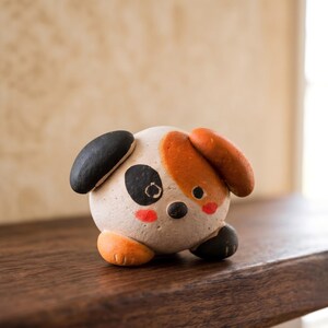 May include: A handcrafted ceramic dog figurine with a round body. The dog has black and orange ears, a black eye patch, and red cheeks. The figurine is sitting on a wooden surface.