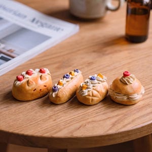 May include: Four miniature pastries sit on a wooden table. The pastries include a cat-shaped bun with red berries, an eclair with purple and yellow toppings, and two other buns with cream and fruit toppings. A coffee mug and a bottle are in the background.