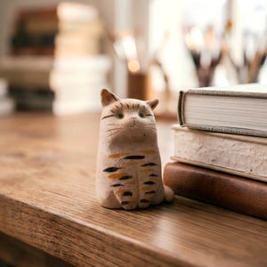 May include: A ceramic cat figurine with a light brown base color and black and orange stripes. The cat has a smiling face and is sitting on a wooden surface. Books are stacked in the background.