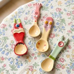 May include: Four whimsical ceramic spoons with cartoon monster designs. The spoons have yellow bowls and colorful handles in red, pink, and green. The spoons are decorated with eyes, mouths, and other playful features. The spoons are arranged on a floral patterned tablecloth.