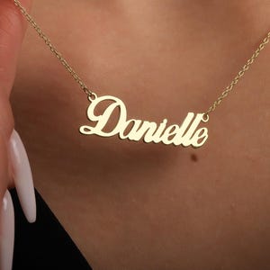 May include: A gold-colored necklace with the name "Danielle" in a cursive font. The necklace is held by a person with long, manicured nails. The chain is delicate and the pendant is centered.