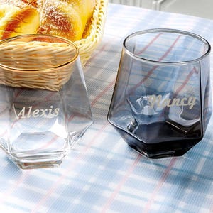 Puede incluir: Dos vasos personalizados, transparentes, de forma geométrica. Un vaso es transparente con el nombre "Alexis" grabado. El otro vaso es gris ahumado con el nombre "Nancy" grabado. Una cesta de pan está en el fondo.
