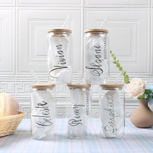 Pode incluir: Um conjunto de cinco copos de vidro transparente com tampas de bambu e canudos transparentes. Cada copo tem um nome em escrita elegante: Vivian, Sloane, Celeste, Remy e Seraphina. A palavra "Bridesmaid" é impressa em um copo. Os copos são preenchidos com papel de seda branco.