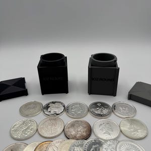 May include: An assortment of silver coins and two black containers labeled "1OZ ROUND" are displayed. The coins feature various designs and are arranged in rows. Two black geometric shapes are on either side of the coins.