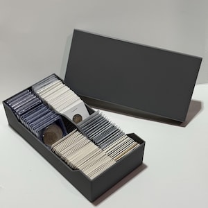 May include: A dark gray coin storage box with a lid, filled with rows of coins in protective sleeves and cardboard holders. The box is open, revealing the organized collection, ideal for coin collectors.