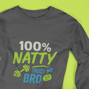 Puede incluir: Camiseta de manga larga gris oscuro con el texto "100% NATTY TRUST ME BRO" en blanco, verde y azul. También incluye un gráfico de mancuernas. La camiseta se muestra sobre un fondo verde brillante.
