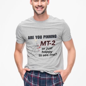 May include: A light gray t-shirt with the text "ARE YOU PINNING MT-2 or just happy to see me?" A drawing of a hand holding a syringe is also on the shirt. The person is wearing plaid pajama pants.