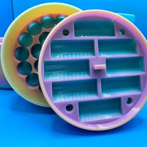 May include: A close-up of two circular, multi-colored 3D-printed objects. One is a container with a series of teal-colored rectangular compartments. The other has a series of teal-colored spheres. A cylindrical container is visible in the background. The objects are on a blue surface.