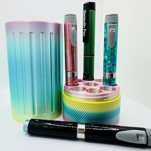 May include: A cylindrical, gradient-colored pen holder with vertical ridges and a separate base featuring multiple circular slots. Three insulin pens with glittery pink, green, and teal finishes are inserted, while a black Gansulin Pen lies in front.