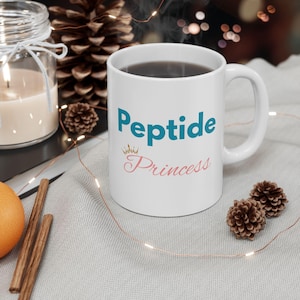 May include: White ceramic mug with the words "Peptide Princess" in teal and pink. The mug has a handle and is filled with a dark liquid. The mug is on a light gray surface with decorative elements.