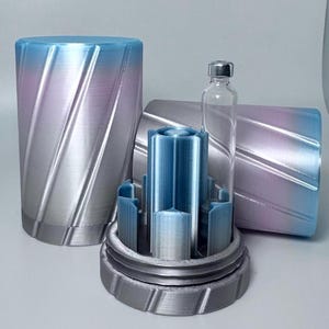 May include: A clear glass vial with a silver cap sits inside a blue and silver 3D-printed holder. The holder is inside a larger, matching container with a gradient of blue, pink, and silver. Two additional containers are visible.