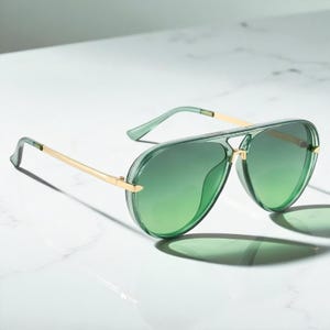 May include: Green aviator sunglasses with gold-colored metal accents. The lenses have a gradient effect, transitioning from a darker green at the top to a lighter shade at the bottom. The sunglasses are set against a white marble surface.