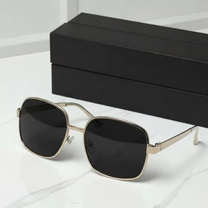May include: Square-framed sunglasses with a gold-colored metal frame and dark lenses. The sunglasses are next to a black rectangular box. The glasses have a modern, minimalist design.