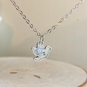 Könnte beinhalten: Eine silberfarbene Halskette mit einem Taubenanhänger. Der Anhänger hat einen weißen Hintergrund mit einem blauen Blumenmuster und einer silbernen Umrandung. Die Kette ist filigran und hängt am Anhänger.
