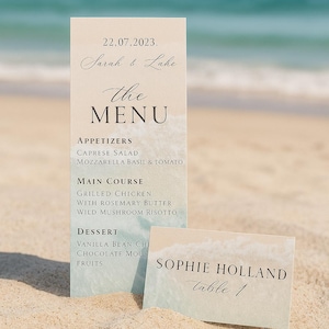 May include: Wedding menu and table card set on a sandy beach. The menu lists appetizers, main courses, and dessert options. The table card displays a name and table number. The background shows the ocean.