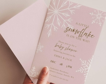 Op de afbeelding: Een lichtroze uitnodigingskaart voor een babyshower met witte sneeuwvlokillustraties en gouden tekst die luidt: A LITTLE snowflake IS ON THE WAY! Please join us for a baby shower honoring parents-to-be EMMA & JACK DEC 18 3 PM.
