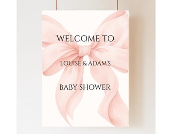 May include: A baby shower sign featuring a large, watercolour-style pink bow. The sign reads "WELCOME TO LOUISE & ADAM'S BABY SHOWER" in black text. The background is white, and the sign is suspended by two thin, brown cords.