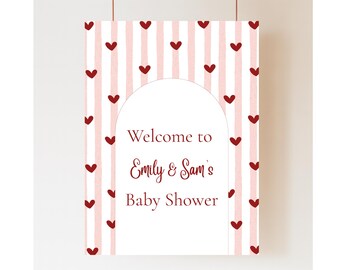 May include: A rectangular sign with a pink and white striped background, adorned with maroon hearts. The sign features an arched white space with the text "Welcome to Emily & Sam's Baby Shower" in maroon script.