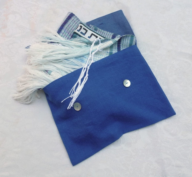 May include: A blue fabric pouch with two buttons and a white fringe. The pouch is open and a patterned prayer shawl with Hebrew writing is inside.