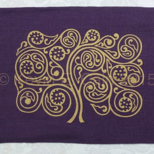 May include: A purple linen placemat with a gold-colored tree design. The tree is stylized with intricate swirls and leaves.