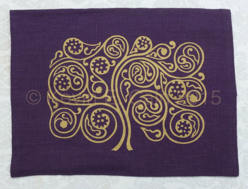 May include: A purple linen placemat with a gold-colored tree design. The tree is stylized with intricate swirls and leaves.