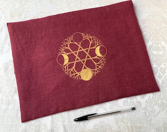 May include: A burgundy-colored pillowcase with a gold-colored, stylized celestial design. The design features a central circle, three crescent moons, and a network of lines and branches. A black and clear pen rests on the surface.