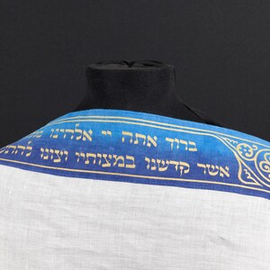 May include: A white prayer shawl with a blue and gold trim. The trim features Hebrew text and a decorative design.