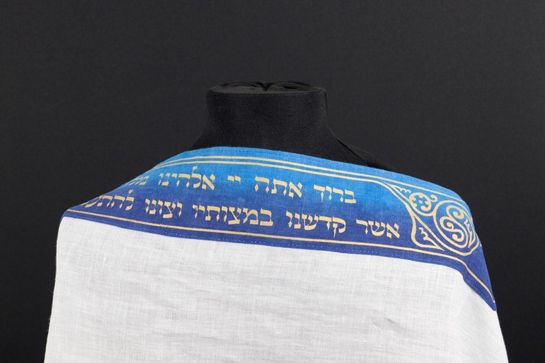 May include: A white prayer shawl with a blue and gold trim. The trim features Hebrew text and a decorative design.