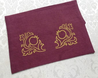 May include: A burgundy linen table runner featuring two gold pomegranate designs. The embroidered pomegranates are framed by ornate, swirling leaf patterns. The runner is rectangular and has a slightly textured appearance, suitable for dining or decorative use.