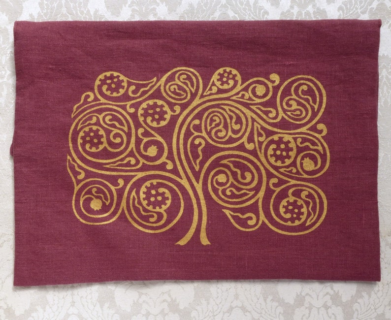 May include: A burgundy linen pillow with a gold embroidered design of a stylized tree with swirling branches and leaves.